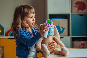 Toddler girl using electronic non-contact thermometer for bunny soft toy in the kids room. Pandemic...