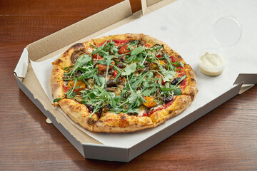 Photo for pizza delivery. veggie pizza with olives, melted cheese, arugula, mushrooms, cherry tomatoes in a cardboard box with side sauce on a wooden table
