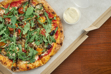 Photo for pizza delivery. veggie pizza with olives, melted cheese, arugula, mushrooms, cherry tomatoes in a cardboard box with side sauce on a wooden table