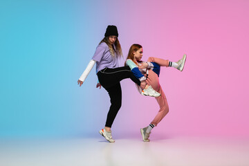 Flexible. Sportive girls dancing hip-hop in stylish clothes on colorful gradient background at...
