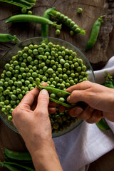 Naklejka na ściany i meble White female hands holding a handful green peas on a wooden table. In the table there are a crystal bowl full of green peas and a linen cloth. Rustic style. Top view.