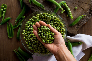 Naklejka na ściany i meble White female hand holding a handful green peas on a wooden table. In the table there are a crystal bowl full of green peas and a linen cloth. Rustic style. Top view.