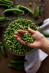 White female hand holding a handful green peas on a wooden table. In the table there are a crystal bowl full of green peas and a linen cloth. Rustic style. Top view.