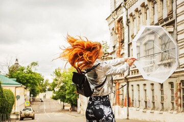 Outdoors lifestyle portrait of ginger hair girl in waterproof silver raincoat walking at downtown. Whirling with an umbrella. Opening transparent umbrella. Rainy weather season concept. Gloomy weather