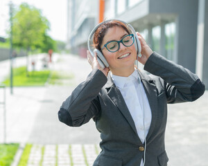 Portrait of a smiling woman in a business suit listening to music in headphones outdoors. Female office employee holds hands on earphones enjoying song and dancing on the street.