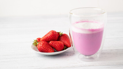 Few ripe strawberries and pink matcha latte with milk in double walled glass on white small plate on wooden table 