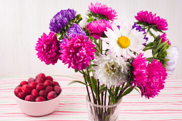 Red plums in pink bowl and glass vase with a bouquet of chrysanthemums on  colorful striped tablecloth.
