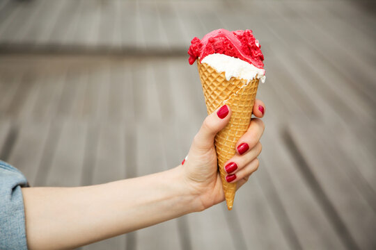 Close up image of woman hand holding fresh waffle cone with vanilla and raspberry ice cream on the wooden background. Walking. Outdoors