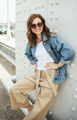 Outdoors fashion portrait of cheerful trendy woman posing on the street. Smiling and walking on the city. Going shopping. Wearing stylish wide jean jacket, beige slacks and sunglasses. close up