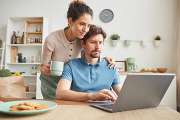 Young sitting at the table in the kitchen and working on laptop with his girlfriend standing nearby and drinknig coffee