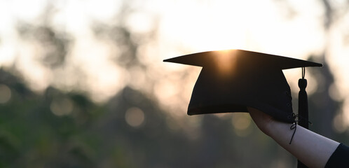 Cropped image of university student hand holding a graduation hat in hand over outdoors with sunset as background. - obrazy, fototapety, plakaty