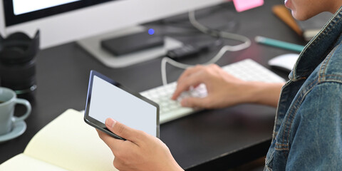 A smart man is holding a white blank screen tablet while typing on a keyboard at the black working desk.