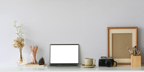 Comfortable workspace that surrounded by white screen computer laptop, wild grass in vase, pine cone, stack of notebook, coffee cup, retro camera, wooden pencil holder and empty picture frame.
