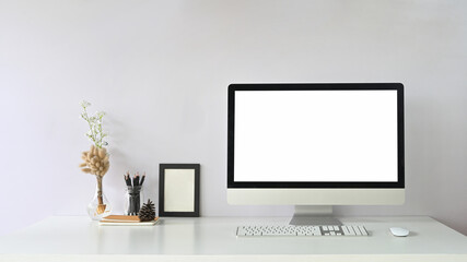 Computer with white blank screen putting on white working desk that surrounded by mouse, keyboard, empty picture frame, pencil holder, pine cone, stack of notebook and plant in jug.