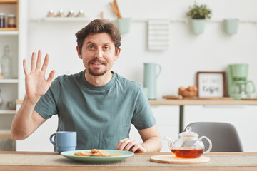 Fototapeta na wymiar Portrait of young man looking at camera and waving his hand while having breakfast in the kitchen
