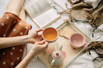 A woman holds a cup of hot tea on the bed at home and reads a book, home leisure in coziness and comfort.