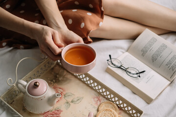 A woman holds a cup of hot tea on the bed at home and reads a book, home leisure in coziness and comfort.