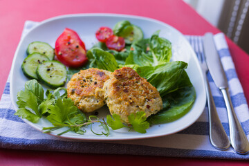 Top view: Delicious Homemade Meatballs or Vegetarian Cutlets, Fresh Vegetables and Green Herbs are on a Plate. It Served on a Red Table with Linen Napkin and Cutlery. 