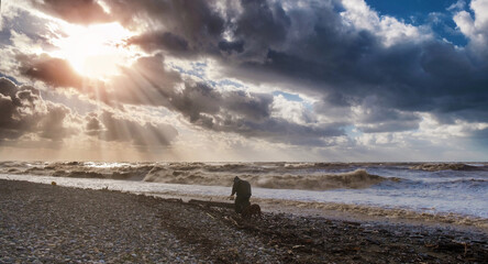  A silhouette of a homeless man with dog walking on the beach with stormy sea and dramatic sunset sky