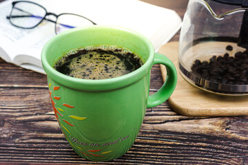 Green cup of coffee with coffee pot and glasses on book in background