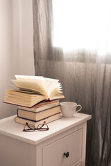 libro abierto sobre una pila de libros, una taza blanca y unas gafas de ver sobre una mesita de...