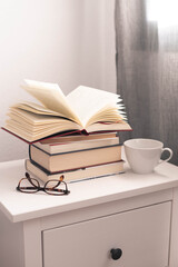libro abierto sobre una pila de libros en una mesita de noche con una taza blanza y unas gafas de...