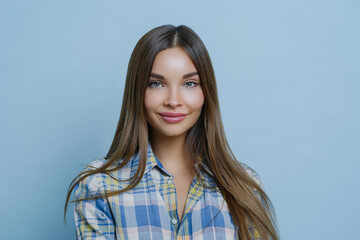 Isolated shot of pretty businesswoman has pleased face expression, tender smile, dressed in casual shirt, has clear skin and attractive facial features, gazes sensually at camera, blue background