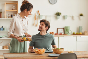 Young wife bringing the dish and serving dinner for her husband who sitting at the table in the kitchen