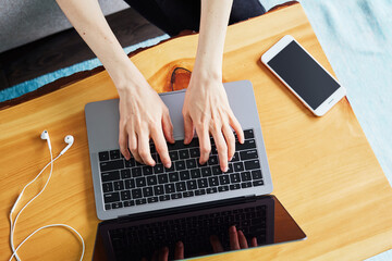 Two hands typing on a laptop with white headphones and a cell phone around, wooden coffee table