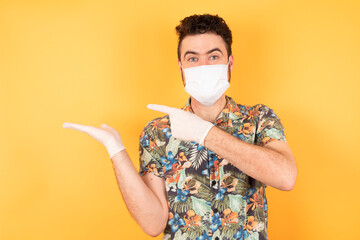 Close up photo of cute young man holding hand showing advertisement wearing hawaiian shirt and medical mask isolated over yellow background