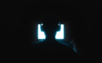 silhouette of a human in headphones sitting in front of a bright screen in the dark