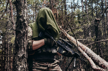 Forest sniper in camouflage and net scarf standing with rifle near the tree.