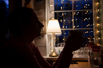 silhouette of a girl with closed eyes holding a white cup with a warm drink and drinking with pleasure. The concept of comfort and warmth. soft focus, background dark window and  garland in blur