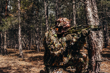 Forest sniper hunter in camouflage leaf and grass suit sitting aiming with crossbow.