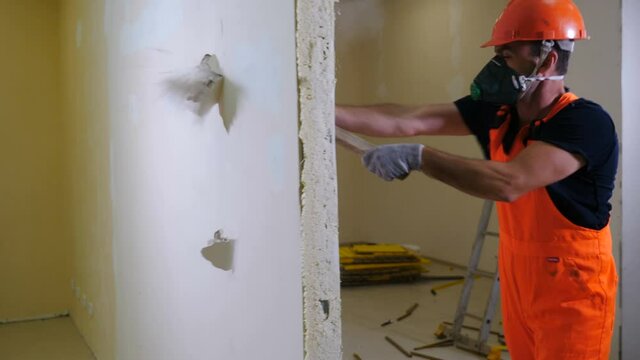Worker in uniform and respirator destroys inner wall sledge hammer. Demolition work and rearrangement concept. Wall made of gypsum cardboard being destroyed. 4 k video