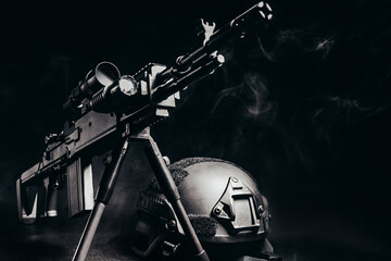 Sniper rifle with scope standing on table with soldier armor helmet on black background with smoke.