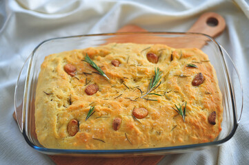Focaccia with garlic and rosemary in a transparent baking dish on a wooden Board on a table covered with a white tablecloth