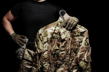 Soldier holding hanger with camouflaged tunic, torso view on black background.