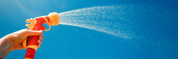A hand holds a yellow hose with a nozzle on a background of blue sky with water splashes. The...