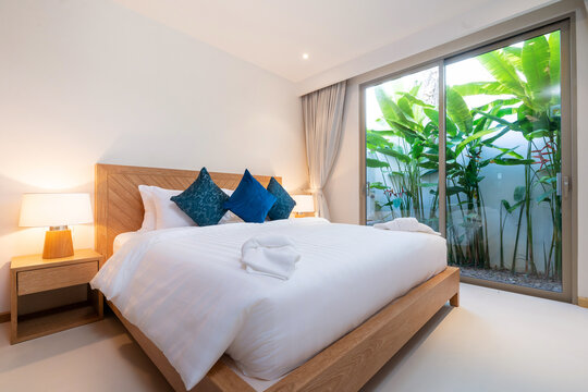 Interior design of bedroom in luxury villa, apartment feature bed, beding, duvet, towels, cushion, bedside table lamp and sluding door with garden view