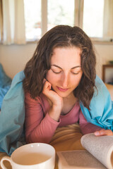 Smiling girl reads a book and drinks coffee while lying in bed.