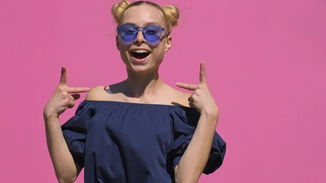 Happy teen blonde girl jumping and waving hands, making funny faces in blue sunny smiles with joy on pink background slow motion in summer. Emotions of people. Positive mood