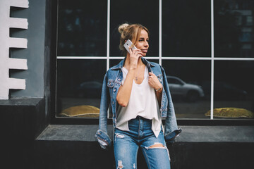 Beautiful hipster girl chatting online standing in urban setting near skyscraper