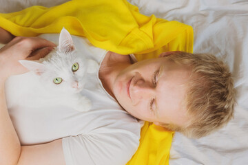 A young blond man is lying on a bed with a white shorthair cat. The concept of pets, weekends, laziness, morning. Photo in bright white and yellow tones.