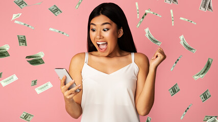 Big Win. Asian Girl Celebrating Victory With Smartphone Under Dollar Money Shower