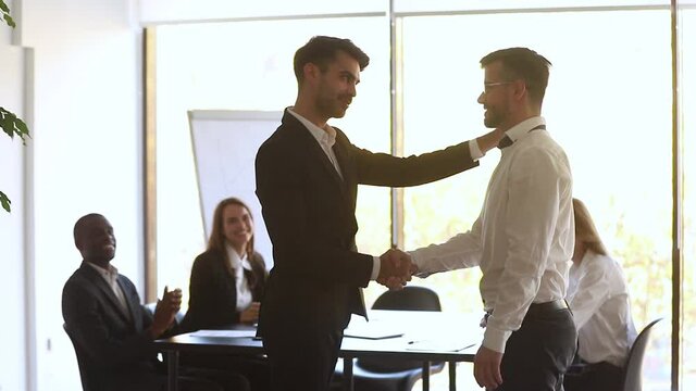 Boss shaking hands welcoming new employee during morning briefing in modern boardroom, diverse staff on background cheers newcomer with applauses, worker of month receive praise gratitude from chief