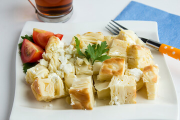 Traditional Turkish Cheesy Water Patty on the white porcelain plate with tomato slices,cutlery set and a cup of tea.