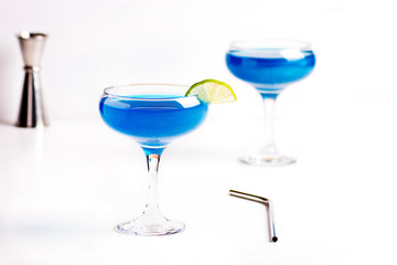 Sparkling blue glitter drinks in lime decorated cocktail glasses are on a white table. Blue lagoon refreshing glitter alcoholic cocktail. Horizontal orientation, selective focus.