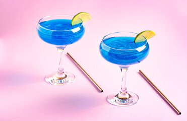 Sparkling blue glitter drinks in cocktail glasses decorated with lime stand on a pink background. Blue lagoon pattern refreshing summer glitter alcoholic cocktail. Horizontal orientation.