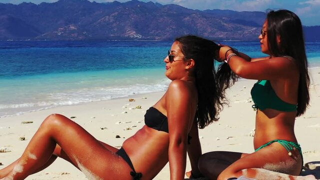 Girl Sunbathing With Her Friend Fixing Her Hair Along The Shore, Tilting Upwards And Exposing The View Of A Mountain Range Over The Calm Sea.